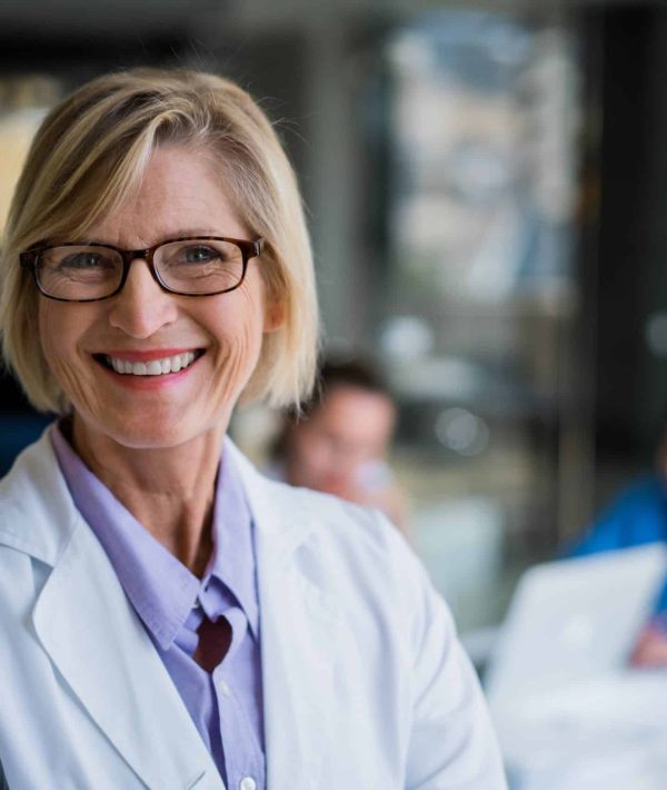 A photo of confident female doctor in hospital. Portrait of happy mature professional wearing lab coat. Smiling expert is with colleagues working in background.